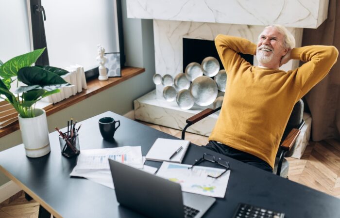 A gray-haired smiling senior man leaned on an armchair holding his hands behind his head while resting after work in a laptop